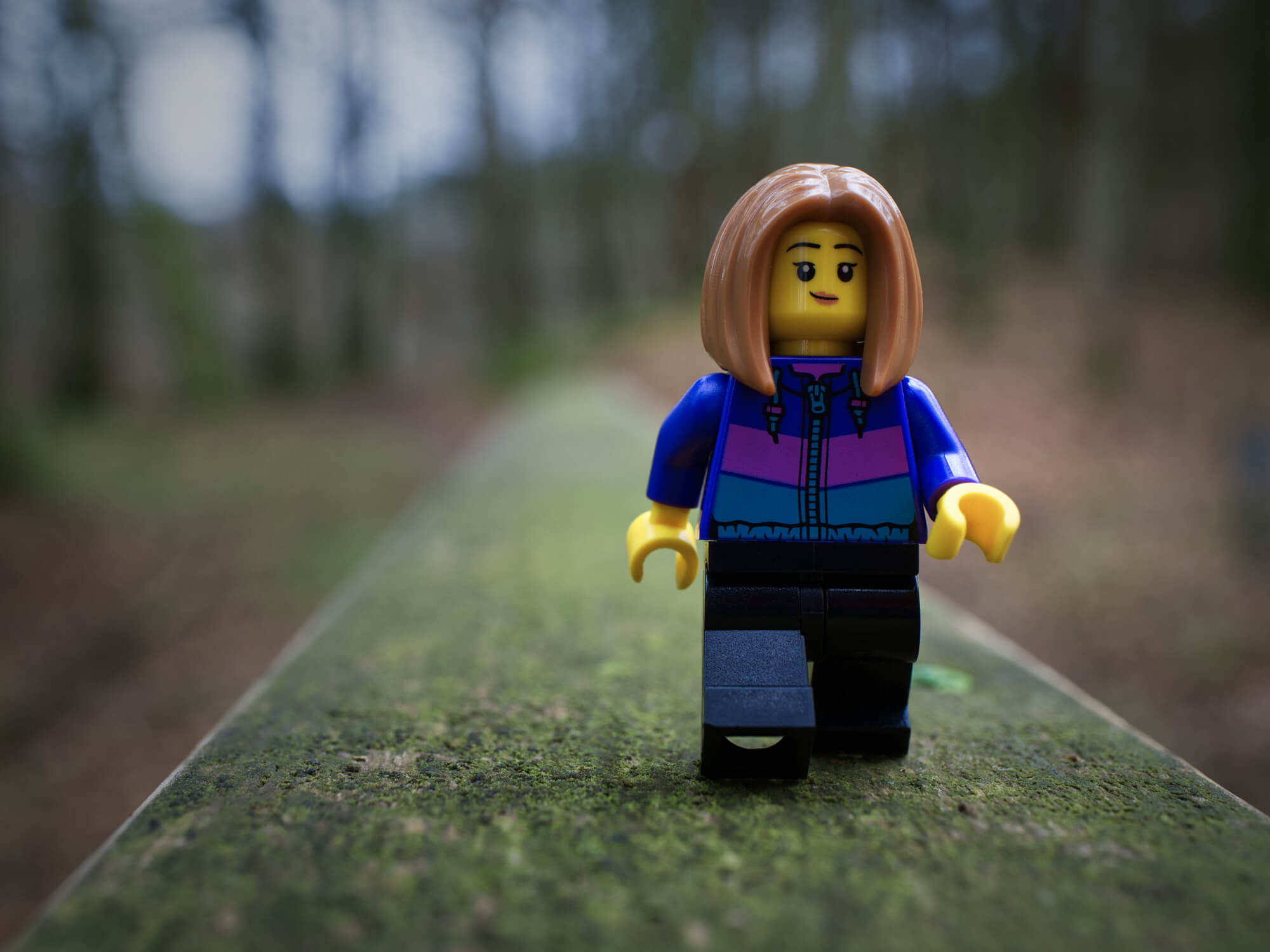 A Lego minifgure walks along a mossy path in a nature scene, with out of focus trees in the background.